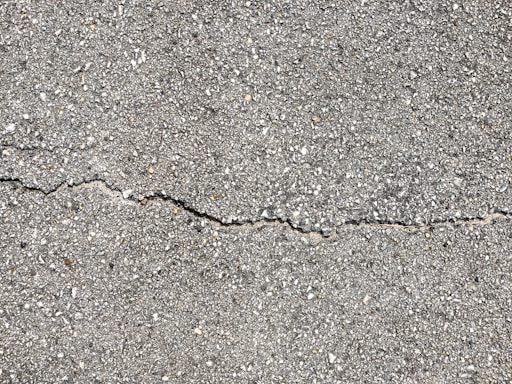 A close-up view of a textured gray concrete surface with a noticeable crack running horizontally across the image. The surface is rough with small pebbles and aggregates visible within the concrete material.