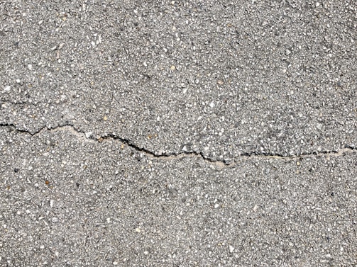 A close-up view of a textured gray concrete surface with a noticeable crack running horizontally across the image. The surface is rough with small pebbles and aggregates visible within the concrete material.