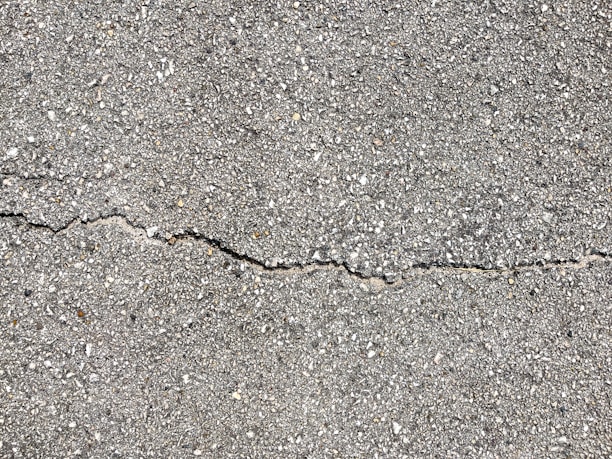 A close-up view of a textured gray concrete surface with a noticeable crack running horizontally across the image. The surface is rough with small pebbles and aggregates visible within the concrete material.