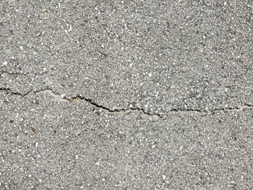 A close-up view of a textured gray concrete surface with a noticeable crack running horizontally across the image. The surface is rough with small pebbles and aggregates visible within the concrete material.