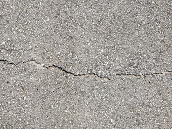A close-up view of a textured gray concrete surface with a noticeable crack running horizontally across the image. The surface is rough with small pebbles and aggregates visible within the concrete material.