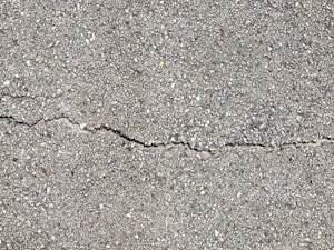A close-up view of a textured gray concrete surface with a noticeable crack running horizontally across the image. The surface is rough with small pebbles and aggregates visible within the concrete material.
