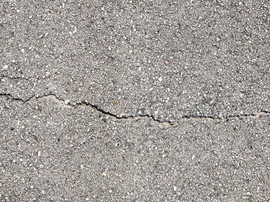 A close-up view of a textured gray concrete surface with a noticeable crack running horizontally across the image. The surface is rough with small pebbles and aggregates visible within the concrete material.
