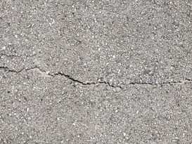 A close-up view of a textured gray concrete surface with a noticeable crack running horizontally across the image. The surface is rough with small pebbles and aggregates visible within the concrete material.