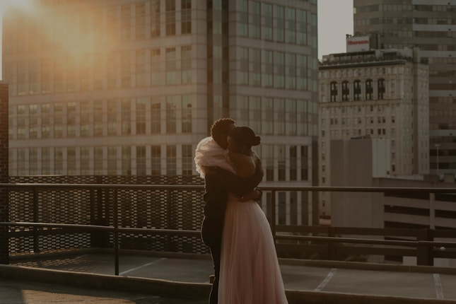 A couple embraces on a rooftop during sunset with tall buildings in the background. The warm golden light of the sun creates a romantic and serene atmosphere.