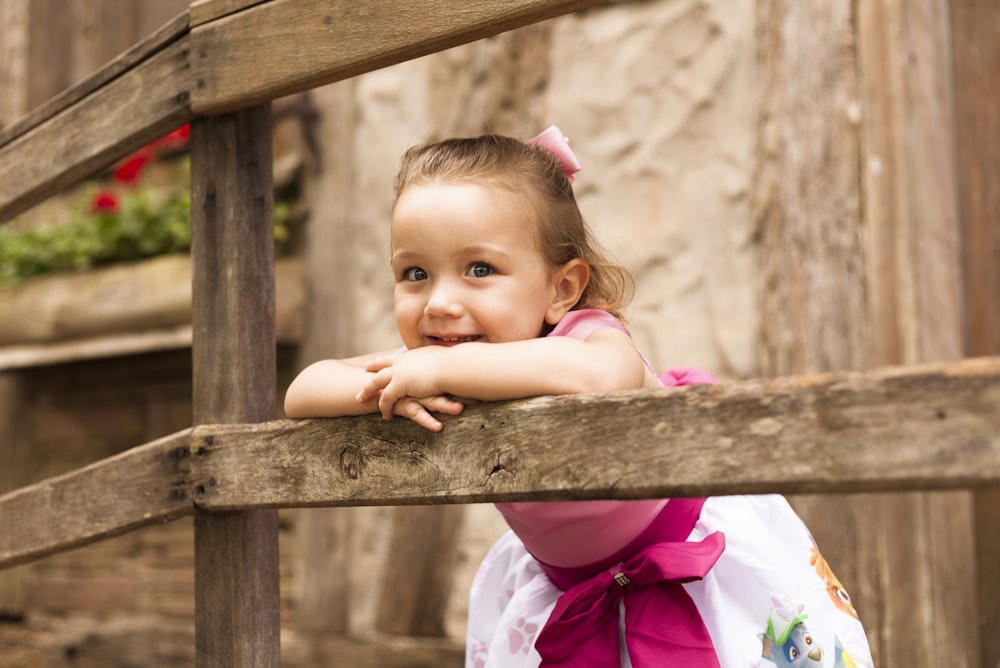 girl in white and pink floral dress leaning on brown wooden fence