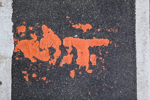 Weathered and chipped orange paint on a rough asphalt surface, flanked by two white lines running parallel to the painted patch.