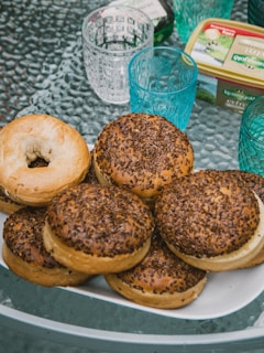 A plate filled with several bagels, some plain and others topped with seeds, sits on a textured glass table. Surrounding the plate are blue glass cups with decorative patterns and a container of Kerrygold butter, adding to the breakfast setting.