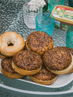 A plate filled with several bagels, some plain and others topped with seeds, sits on a textured glass table. Surrounding the plate are blue glass cups with decorative patterns and a container of Kerrygold butter, adding to the breakfast setting.