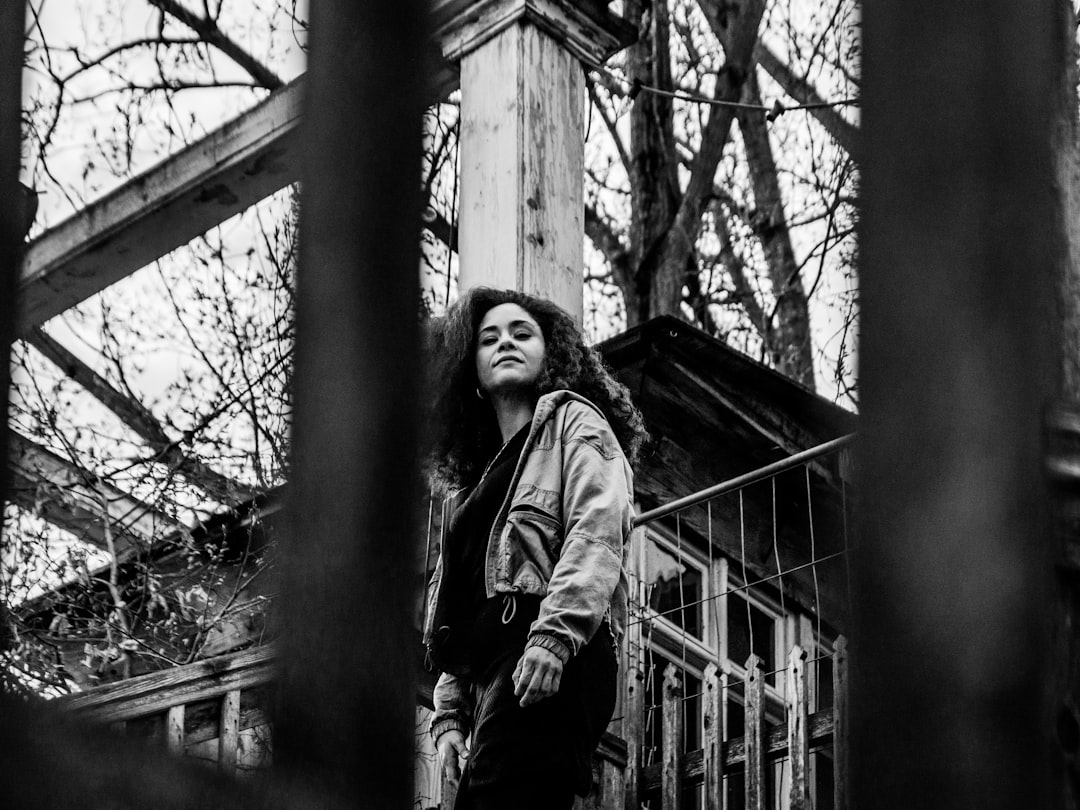 grayscale photo of woman in jacket standing on wooden bridge
