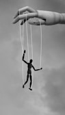 A large wooden hand manipulates a small human puppet using strings. The puppet appears to be suspended in mid-air against a cloudy sky, suggesting themes of control and manipulation.