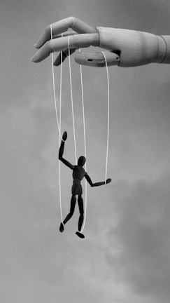 A large wooden hand manipulates a small human puppet using strings. The puppet appears to be suspended in mid-air against a cloudy sky, suggesting themes of control and manipulation.