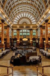 A grand, opulent interior space with a high vaulted ceiling detailed with intricate designs and patterns. The room features tall columns with decorative capitals and large arched windows with stained glass. The floor is patterned with geometric designs, and there are several dining tables set with white tablecloths arranged throughout the space. In the center of the room is a large, ornate chandelier. The atmosphere conveys a sense of historic elegance and luxury.