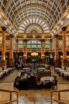 A grand, opulent interior space with a high vaulted ceiling detailed with intricate designs and patterns. The room features tall columns with decorative capitals and large arched windows with stained glass. The floor is patterned with geometric designs, and there are several dining tables set with white tablecloths arranged throughout the space. In the center of the room is a large, ornate chandelier. The atmosphere conveys a sense of historic elegance and luxury.