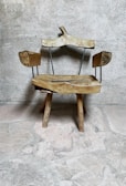 A rustic wooden chair with an uneven, natural finish sits against a textured stone wall. The chair features wooden arms and a backrest supported by thin metal rods, adding a minimalist design touch. The rough stone floor complements the chair's organic and rustic appearance.
