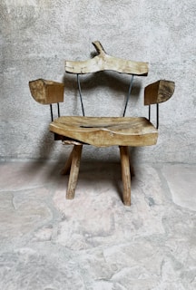 A rustic wooden chair with an uneven, natural finish sits against a textured stone wall. The chair features wooden arms and a backrest supported by thin metal rods, adding a minimalist design touch. The rough stone floor complements the chair's organic and rustic appearance.