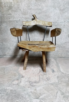A rustic wooden chair with an uneven, natural finish sits against a textured stone wall. The chair features wooden arms and a backrest supported by thin metal rods, adding a minimalist design touch. The rough stone floor complements the chair's organic and rustic appearance.