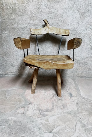 A rustic wooden chair with an uneven, natural finish sits against a textured stone wall. The chair features wooden arms and a backrest supported by thin metal rods, adding a minimalist design touch. The rough stone floor complements the chair's organic and rustic appearance.