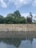 A stone wall stands above a reflective body of water with a layer of smooth pebbles beneath the surface. Behind the wall, lush green trees rise against a sky filled with soft, scattered clouds.