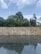 A stone wall stands above a reflective body of water with a layer of smooth pebbles beneath the surface. Behind the wall, lush green trees rise against a sky filled with soft, scattered clouds.