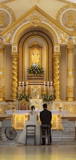 A couple in wedding attire is kneeling before an ornate and gilded church altar. The altar is adorned with white flowers, tall candles, and a central religious icon. The architecture features intricate details and gold accents, showcasing a blend of classical and religious elements.