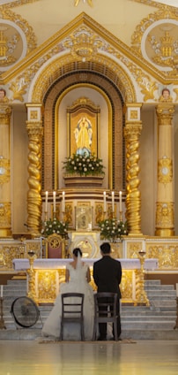 A couple in wedding attire is kneeling before an ornate and gilded church altar. The altar is adorned with white flowers, tall candles, and a central religious icon. The architecture features intricate details and gold accents, showcasing a blend of classical and religious elements.