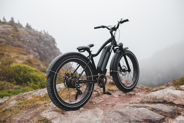A sleek, black electric bicycle is parked on a rocky path surrounded by a misty, mountainous landscape. Sparse vegetation can be seen growing between the rocks, and distant hills are barely visible through the fog.
