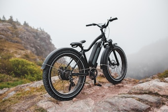 A sleek, black electric bicycle is parked on a rocky path surrounded by a misty, mountainous landscape. Sparse vegetation can be seen growing between the rocks, and distant hills are barely visible through the fog.