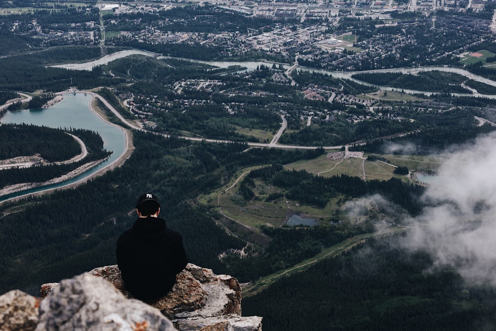 Mann in schwarzer Jacke sitzt tagsüber auf Felsen und schaut auf die Stadt