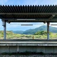 A train platform under a corrugated metal roof is set against a backdrop of lush green hills and mountains under a clear blue sky. The platform is empty, with a strip of yellow tactile paving along its edge. Grass and shrubs grow around the area, adding to the serene and natural setting.
