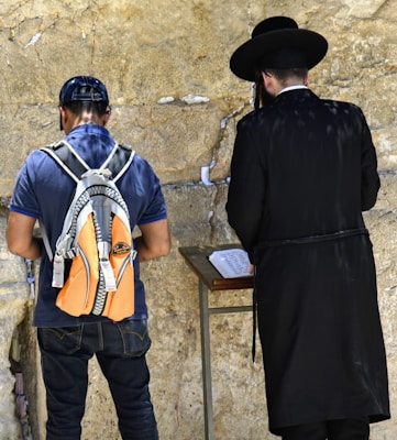 Two people are standing in front of a stone wall, engaged in prayer or contemplation. One person is wearing a black coat and hat, while the other wears a casual outfit with a cap and a colorful backpack.