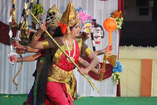 Several individuals dressed in traditional attire are performing a dance or theatrical act. They are holding various weapons and props, wearing vibrant and ornate costumes, including headdresses. The background features decorative elements like flowers and drapery.