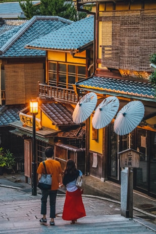 A couple is seen walking hand in hand down a narrow stone pathway lined with traditional Japanese buildings. The architecture features wooden facades and blue tiled roofs. Paper lanterns provide warm lighting, enhancing the tranquil ambiance. Decorative white umbrellas are mounted on the exterior of one building.