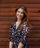 A woman with long brown hair is smiling while standing in front of a wooden wall. She is wearing a dark floral dress with various colors.