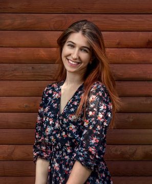 A woman with long brown hair is smiling while standing in front of a wooden wall. She is wearing a dark floral dress with various colors.