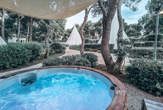 A hot tub with bubbling water is situated outdoors, surrounded by greenery and trees. In the background, several teepee tents are set amongst the trees. The scene is under a large canopy that provides partial shade.