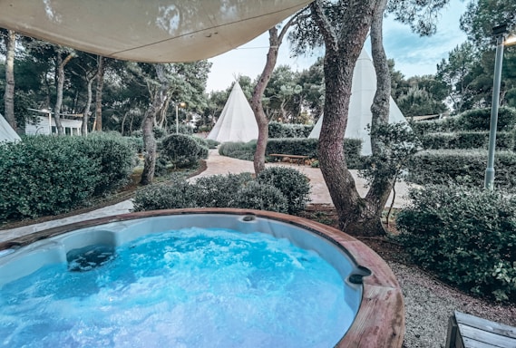 A hot tub with bubbling water is situated outdoors, surrounded by greenery and trees. In the background, several teepee tents are set amongst the trees. The scene is under a large canopy that provides partial shade.
