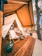 A cozy glamping setup with a wooden deck featuring a large canvas tent. On the deck, there are two wooden lounge chairs with white fabric seats and a small side table between them holding decorative plants and articles. The surrounding area includes large trees and a clear sky, suggesting an outdoor, natural environment.