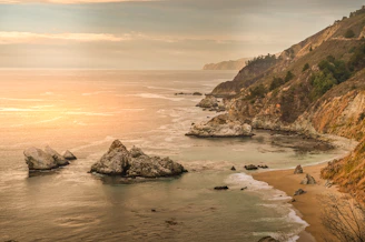 A serene sunrise over a rocky shoreline with gentle waves lapping the coast.
