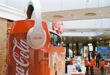 A large Coca-Cola can replica with oversized white headphones is prominently displayed in a modern indoor setting, likely a shopping mall. In the background, several people, some wearing masks, are seen interacting with different exhibits and displays. The environment has a commercial and lively atmosphere with signs and posters visible.