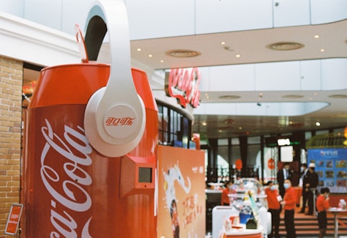 A large Coca-Cola can replica with oversized white headphones is prominently displayed in a modern indoor setting, likely a shopping mall. In the background, several people, some wearing masks, are seen interacting with different exhibits and displays. The environment has a commercial and lively atmosphere with signs and posters visible.
