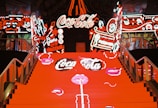 A vibrant staircase with bright red steps leading up, covered in neon signs and logos. Surrounding walls are adorned with colorful neon graphics featuring white and red designs, along with stylized illustrations and text.