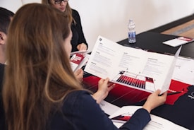 A group of people is sitting around a table engaged in a discussion. One person, with long hair, is holding a brochure or pamphlet featuring text, images, and charts. The setting appears to be a meeting room with documents spread out on the table. Items such as a water bottle and pens are visible.