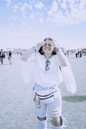 A person stands in a sunny outdoor setting, laughing with their hands covering their eyes. They are wearing a white top with flowing sleeves, ripped jeans, and a belt bag. The sky is blue with scattered clouds, and there are other people and tents visible in the background.