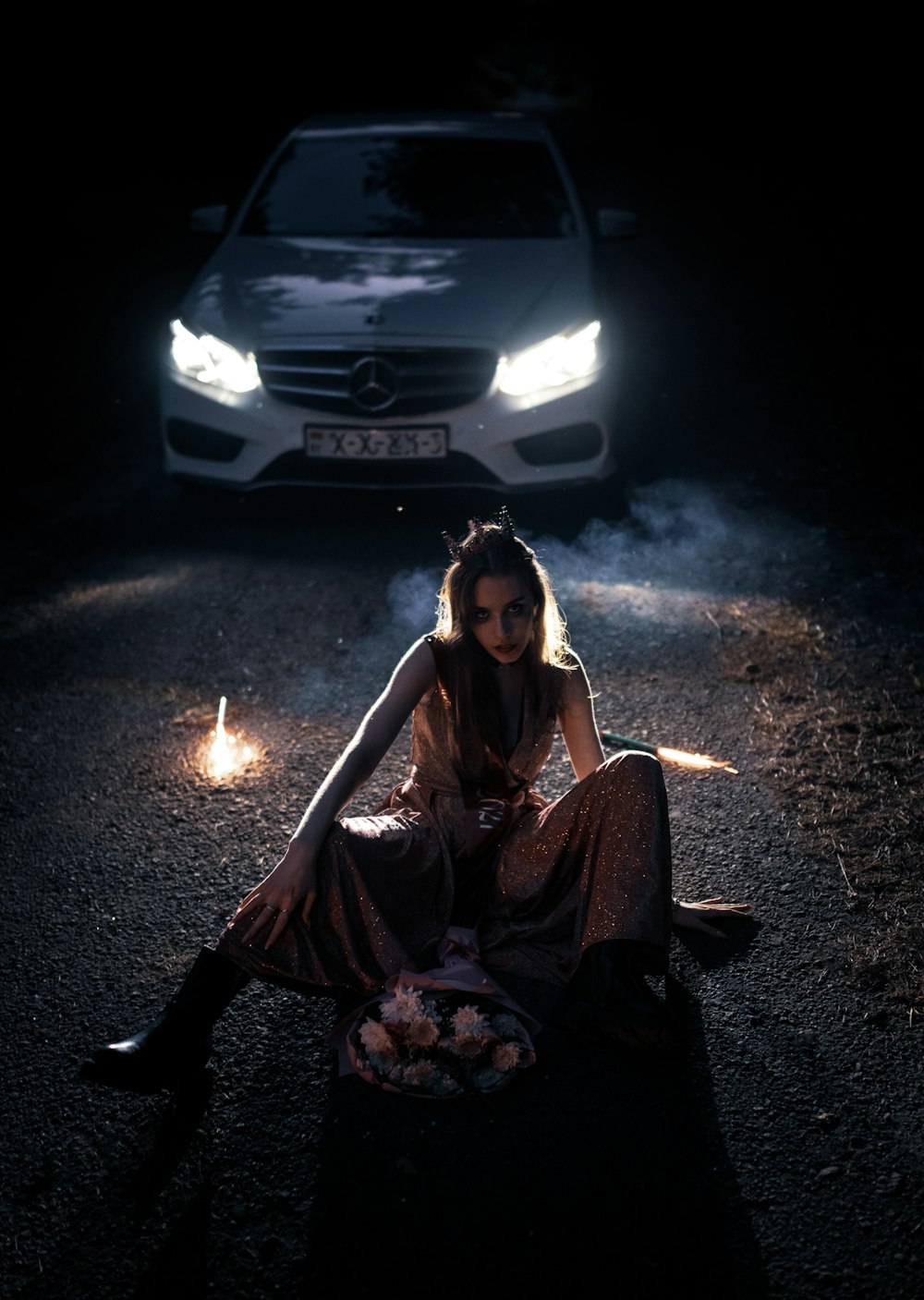 woman in brown long sleeve shirt sitting on ground beside white car during nighttime