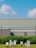 A modern architectural scene with a metal building in the background, featuring a flat roof and a paneled facade. In the foreground, there are several metallic sculptures arranged in a grassy area. Tall, lush green grass borders the sculptures, contrasting with the clear, vibrant blue sky overhead, punctuated by fluffy white clouds.