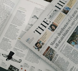 A section of a newspaper is placed over an open magazine. The newspaper displays a headline about the distribution of ex gratia finance and includes several smaller images and text flags. The magazine features articles and highlights, including historical and political content, with images and diagrams accompanying the text. The overall composition suggests a mix of current news and historical retrospectives.