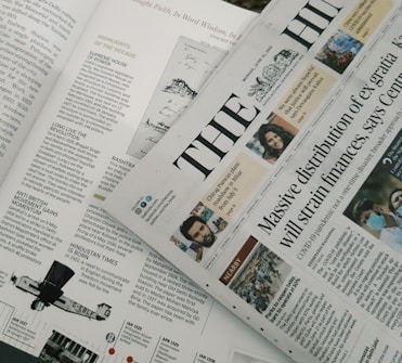A section of a newspaper is placed over an open magazine. The newspaper displays a headline about the distribution of ex gratia finance and includes several smaller images and text flags. The magazine features articles and highlights, including historical and political content, with images and diagrams accompanying the text. The overall composition suggests a mix of current news and historical retrospectives.