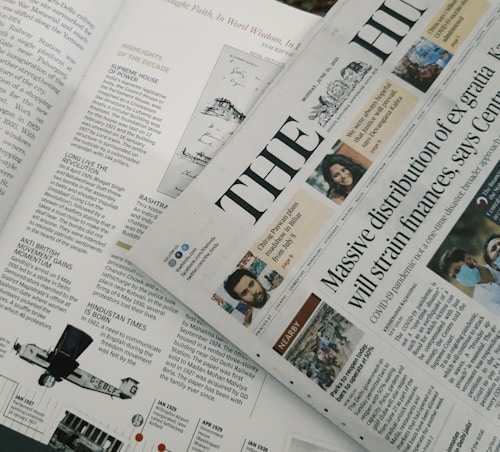 A section of a newspaper is placed over an open magazine. The newspaper displays a headline about the distribution of ex gratia finance and includes several smaller images and text flags. The magazine features articles and highlights, including historical and political content, with images and diagrams accompanying the text. The overall composition suggests a mix of current news and historical retrospectives.