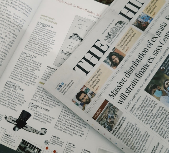 A section of a newspaper is placed over an open magazine. The newspaper displays a headline about the distribution of ex gratia finance and includes several smaller images and text flags. The magazine features articles and highlights, including historical and political content, with images and diagrams accompanying the text. The overall composition suggests a mix of current news and historical retrospectives.